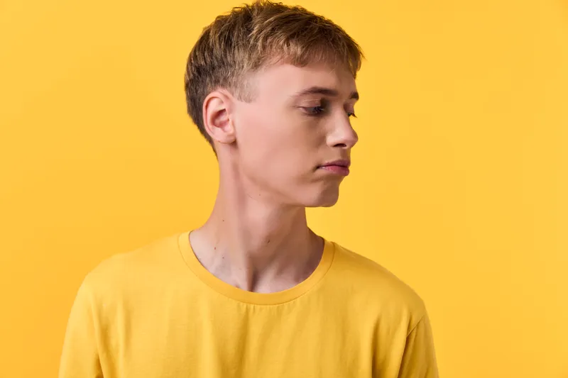 Young man in a yellow shirt turns slightly to the side, clean studio vibe with a bright yellow backdrop, conveying simplicity, focus, and contemporary style.