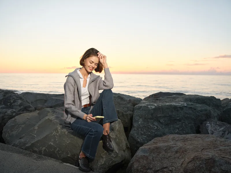 A cheerful young woman sits on rugged rocks beside the sea at sunset, texting on a mobile phone with a bright smile, casual clothing, and a relaxed mood.