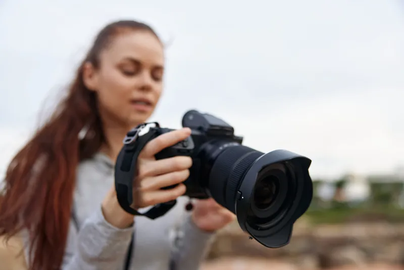 Young woman with long brown hair holding a camera outdoors, showcasing passion for photography, dressed casually in a hoodie, creating a creative atmosphere