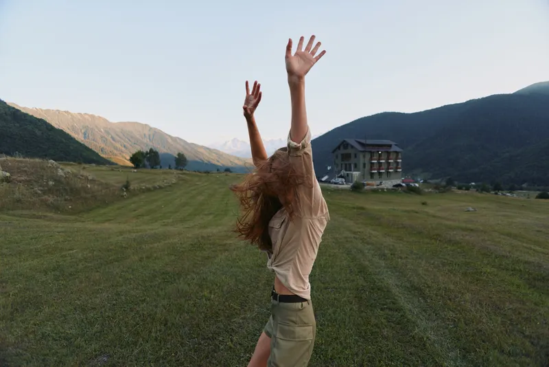 Young woman lifestyle enjoying freedom outdoors in green field with mountains and house in background raising hands up with joyful mood during sunny day.