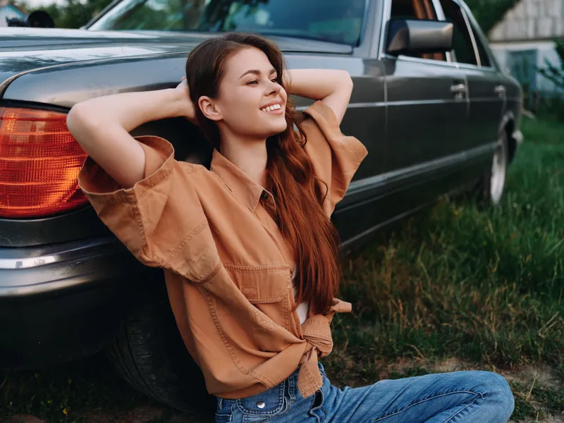 Young woman with long hair relaxing outdoors near a vintage car, wearing a casual brown shirt and jeans, exuding joy amidst a lush green background.