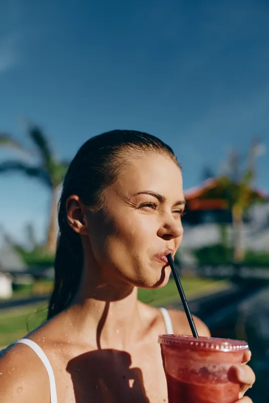 Smiling woman sipping a smoothie outdoors, sunny poolside scene with tropical greenery, vibrant wellness lifestyle and self care moment, refreshing thirst quencher with bright, warm lighting
