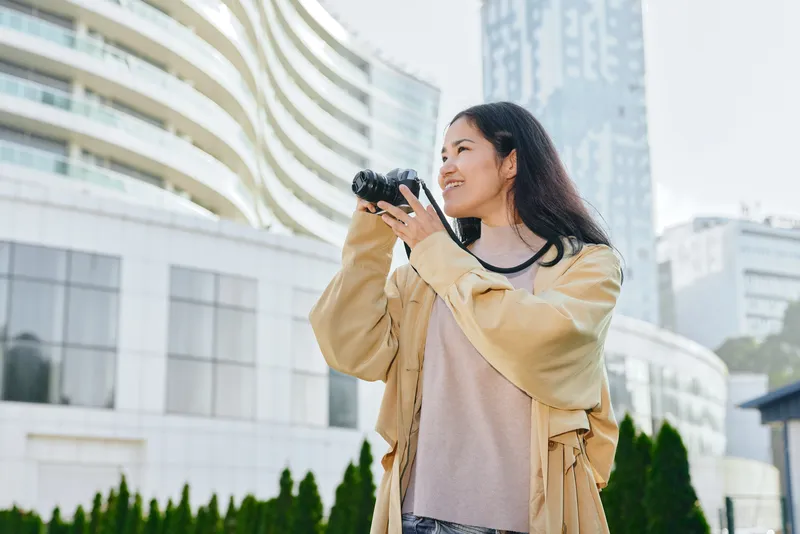Camera in hand, photographer smiles while capturing the city skyline from a sunny outdoors setting, urban street scene, travel vibe, landscape focus, casual attire, vibrant mood, authentic street