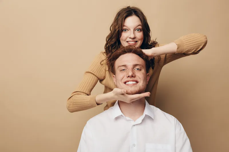 Happy young couple poses together on beige background, woman playfully framing man's face with hands. Smiling man and woman show joyful mood and close relationship concept.