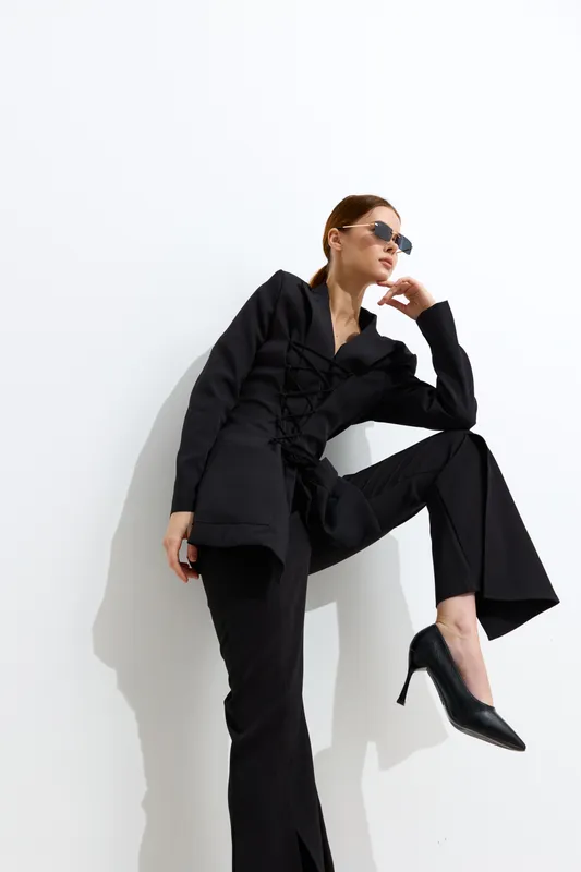 Fashion woman dressed in black stylish outfit with sunglasses poses against white background, showcasing modern chic, high heels, and confident attitude in minimalist setting.