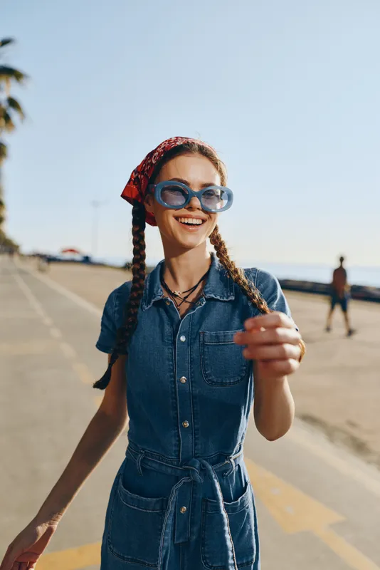 Lifestyle woman in modern boho-western denim street style smiling outdoors during sunny day, wearing blue sunglasses and red bandana, film color vibe