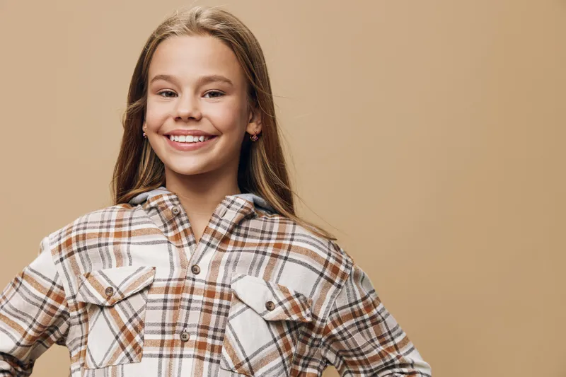 Girl smiling and posing with hands on hips in warm plaid shirt against beige background. Child with long hair and joyful expression in casual autumn outfit for portrait.