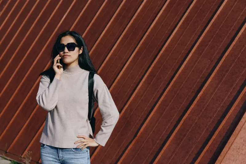 Woman in a chunky knit sweater and sunglasses stands against a diagonal wooden wall, talking on a phone with a backpack, showcasing casual urban style and everyday communication