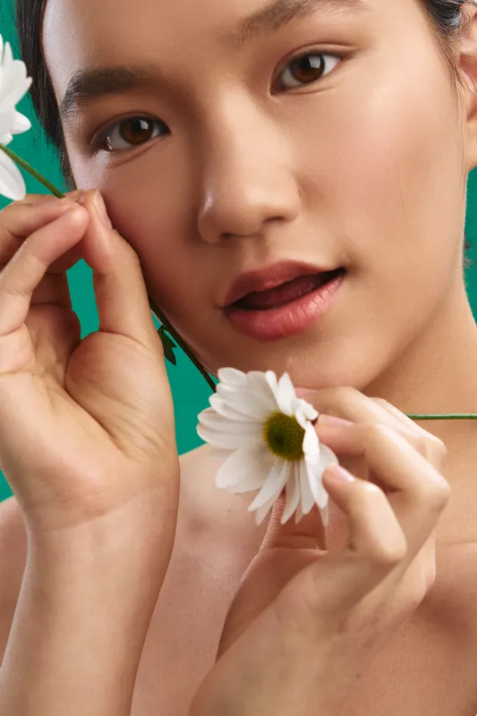 Asian woman gently holds white daisies against a vivid backdrop, focusing on delicate petals and smooth skin, conveying calm beauty and graceful expression.