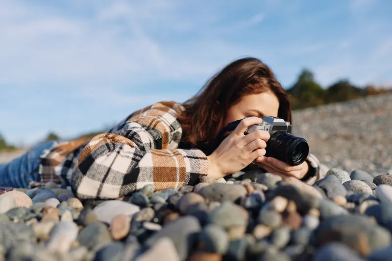 Woman photographer lying on rocky beach, aiming a camera at the horizon for landscape shots, wearing a plaid jacket, clear sky, focused expression, and a casual outdoor photography session.