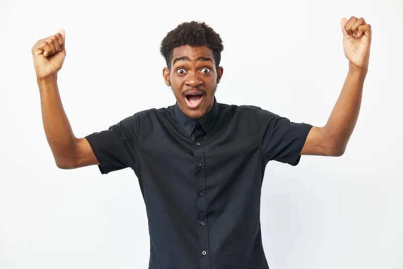 Excited young black man celebrating success with raised hands, conveying joy and enthusiasm in a bright and cheerful atmosphere. Perfect for inspirational themes.