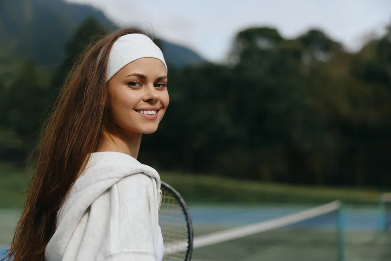 Young female tennis player smiling confidently on the court, wearing a headband and a hoodie, showcasing a healthy athletic lifestyle