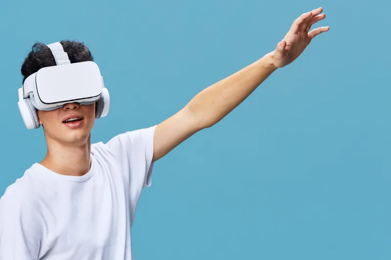 Young man wearing virtual reality headset enjoys immersive digital experience, raising arm against blue wall, technology, innovation, entertainment, modern lifestyle.