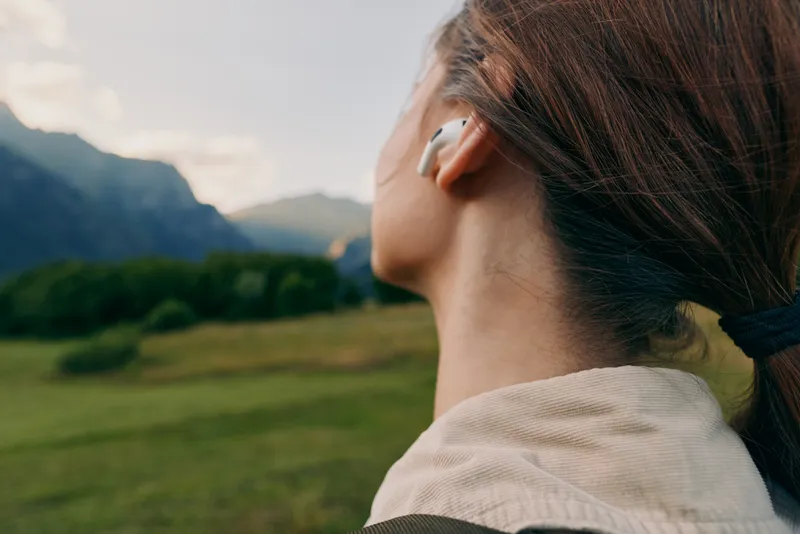 Young woman with brunette hair tied in a ponytail listens to music with wireless earbuds while enjoying a peaceful outdoor landscape with mountains and green fields.