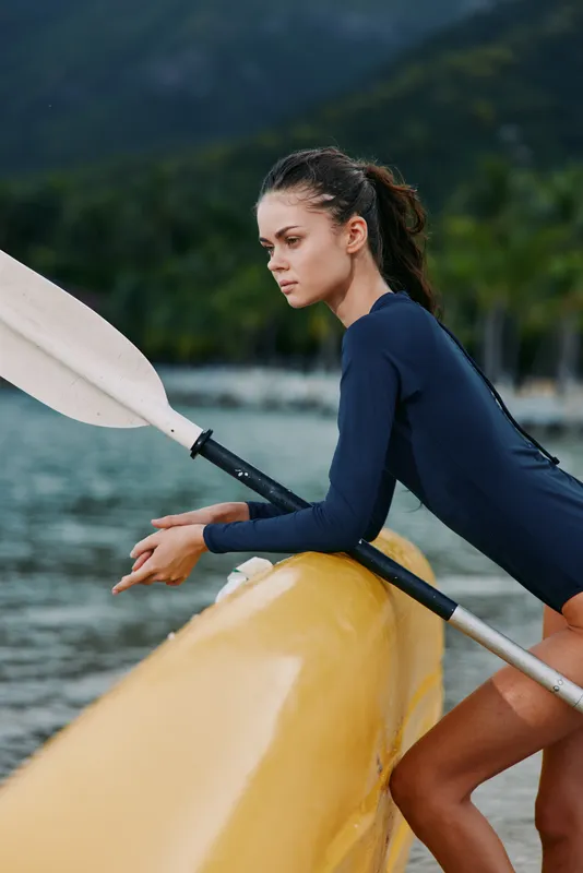Female athlete leaning on a bright yellow canoe, gripping a paddle, focused and poised by calm water, athletic silhouette, outdoor sport scene with distant hills.