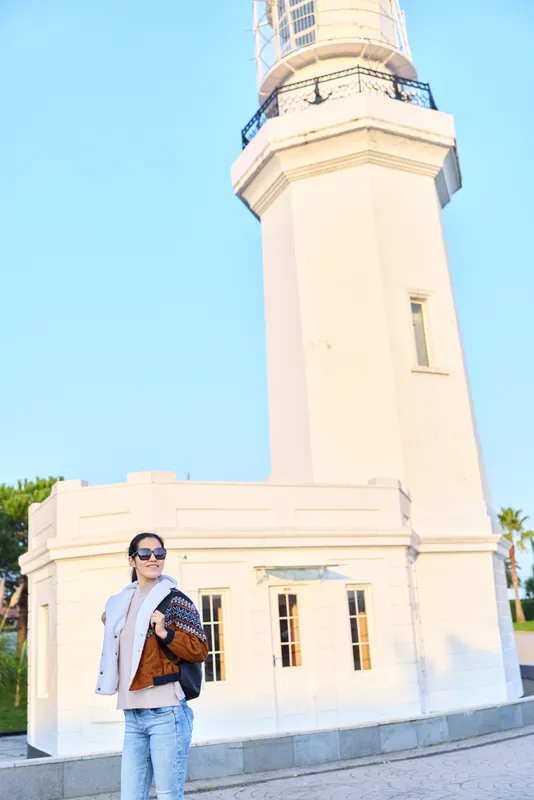 Lighthouse exterior with a stylish woman standing in front, wearing sunglasses, denim jacket, scarf, and a bag, posing on a bright sunny day under a clear blue sky, with architectural surroundings