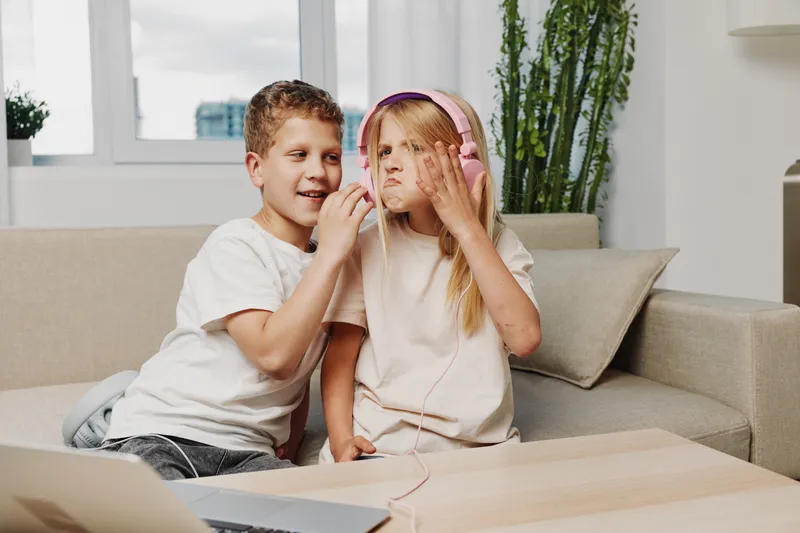 Two children share headphones while sitting on a couch in a bright living room. The boy smiles as the girl reacts with a funny expression, creating a playful moment.
