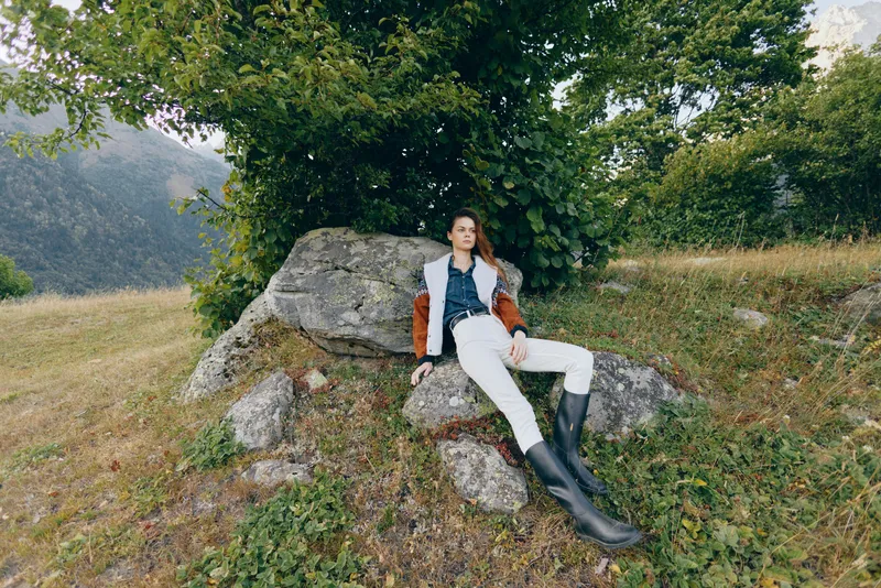 Woman in nature by mountains sitting on a rock under a tree outdoors, fashion model in jacket, white pants and tall boots relaxed in a meadow landscape for travel and leisure