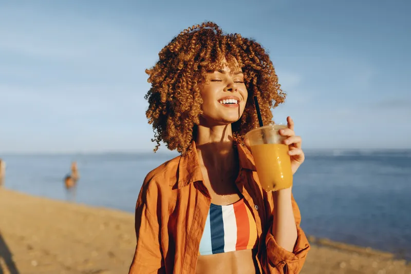 Young woman with curly hair enjoys a fresh orange juice on the beach under clear sky, wearing a striped swimsuit and an open shirt, smiling with closed eyes in warm sunlight.