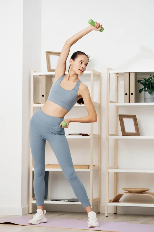 Woman in blue athletic wear performs a dumbbell shoulder press at home, showcasing fitness, balance and focus in a bright living space with shelves and plants.