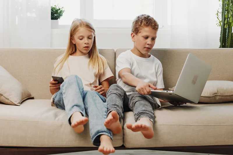 Two children sitting barefoot on a beige sofa using laptop and remote control in a bright living room. Concept of technology, childhood, and casual home life.