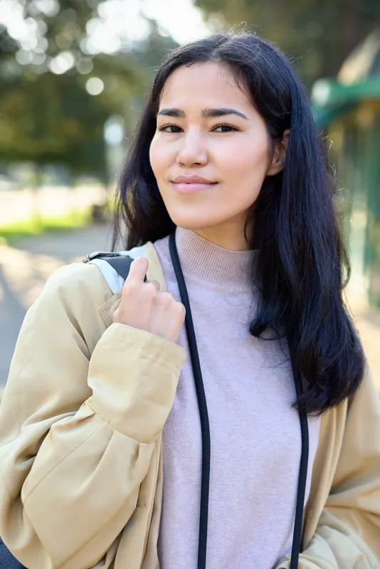 Woman in casual beige jacket and soft knit sweater stands outdoors in a park, carrying a shoulder bag, calm expression, natural lighting, candid lifestyle vibe with warm tones