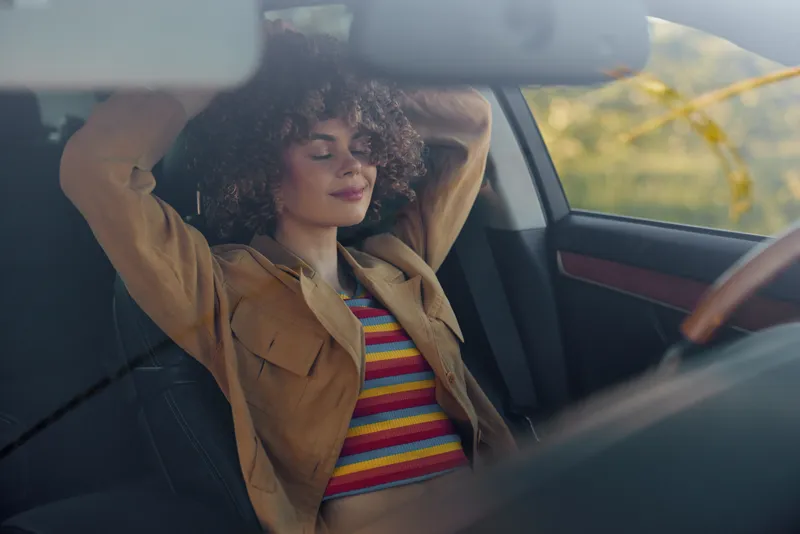 Woman with curly hair sits in a car, arms raised behind her head, wearing striped shirt and tan jacket, capturing a casual lifestyle moment with retro vintage film color vibes.