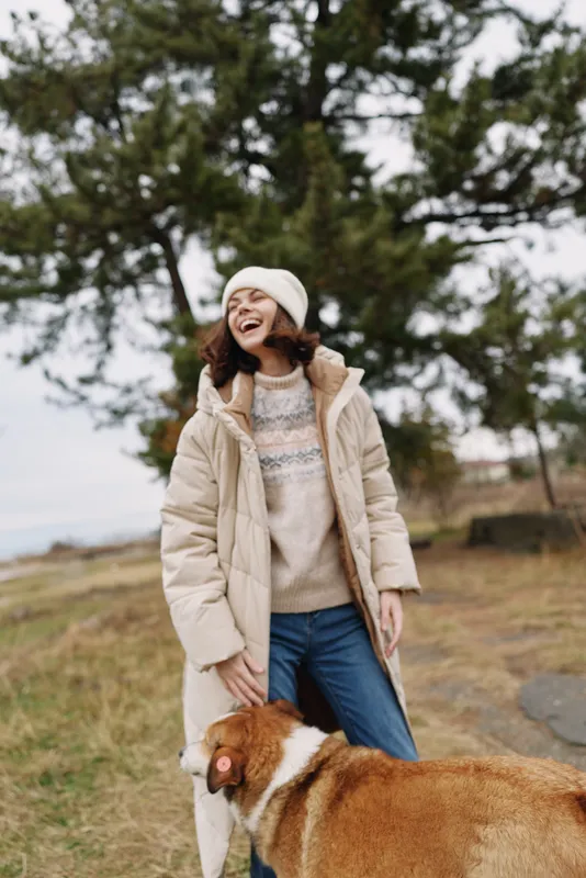 Woman in a thick winter coat, beanie and sweater laughs with a large dog outdoors in a park, trees and open scenery in the background, a casual lifestyle moment captured in chilly weather with