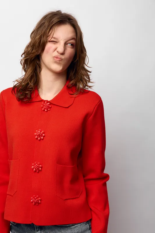 Young woman in red sweater makes funny face with squinted eyes and pursed lips against white background. Casual style, playful mood, and expressive portrait of female model.