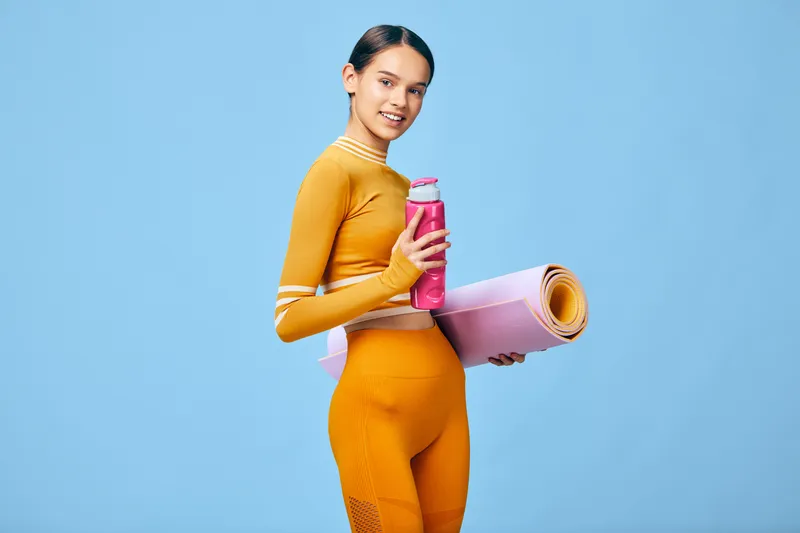 Fitness woman in bright orange active wear stands against a blue backdrop, holding a pink water bottle and a rolled yoga mat, ready for a productive workout and daily wellness routine. Her pose