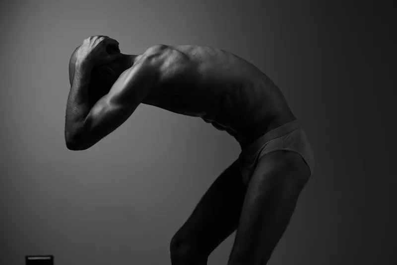 Black and white image of a man expressing deep emotions, showcasing his physical form and vulnerability through dramatic pose and lighting.