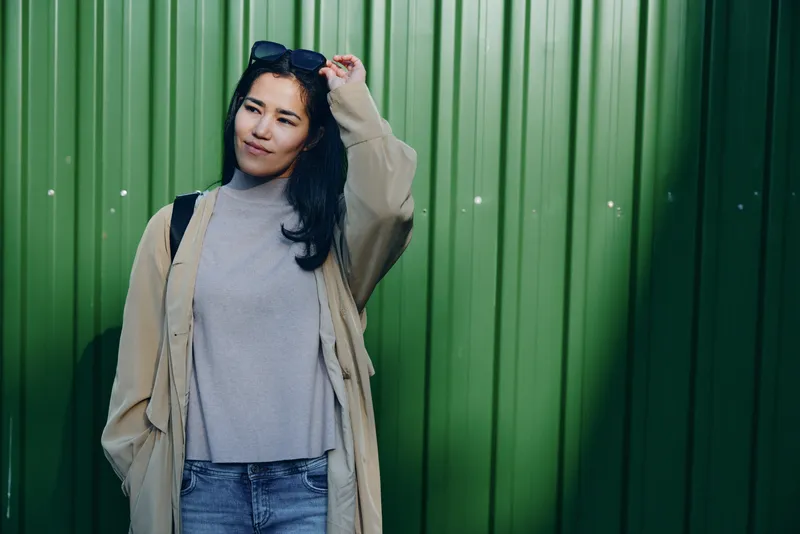 Woman in beige coat and gray sweater stands against a green corrugated wall, casual street fashion, confident pose, outdoor setting with soft light glow.