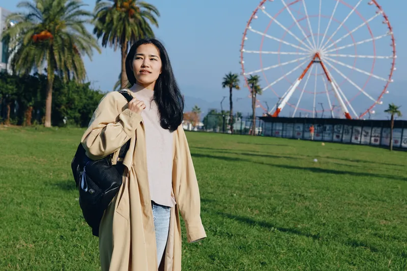 Woman in a light beige coat and white top stands in a sunny park with a large ferris wheel in the background, carrying a backpack for a casual outdoor travel scene.