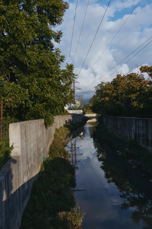 Concrete canal with water reflection, trees and power lines under blue sky and clouds. Photographer shadow on wall, urban nature scene with calm stream and leafy embankment.