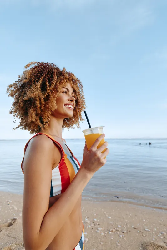 Woman with curly hair smiling profile on sunny beach, holding a cold drink with straw. Ocean and blue sky behind. Summer vacation relaxation, wellness, hydration.