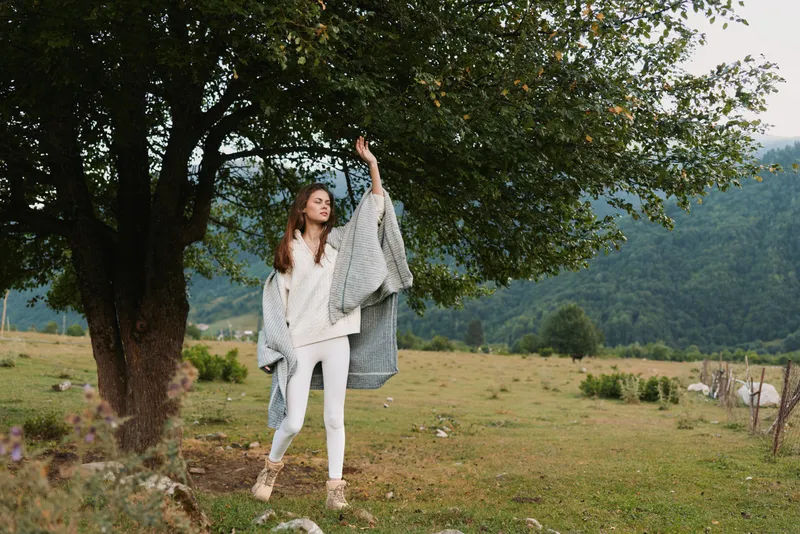 Young woman in white clothes and gray shawl stands near tree in green meadow with mountains on background enjoying nature and peaceful outdoor lifestyle.