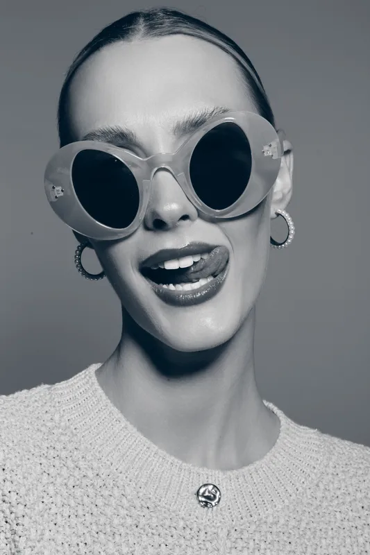Young white woman with pale skin and slicked hair wears oversized round sunglasses and hoop earrings. She smiles playfully sticking out her tongue against neutral background.