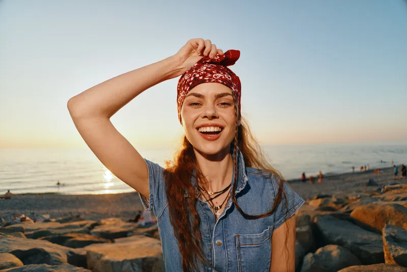 Lifestyle woman in modern boho-western denim street style smiling with a red bandana, natural makeup, and sea sunset background in warm film color tones.