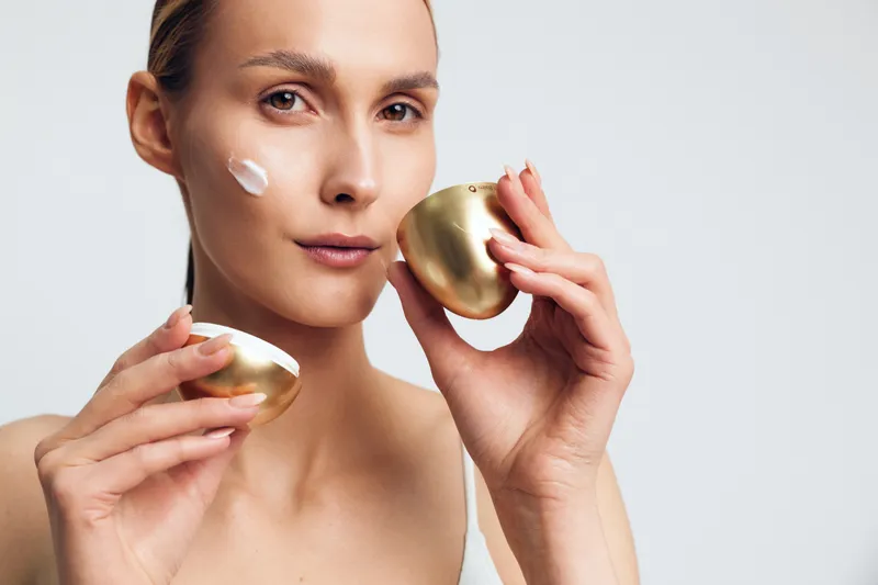 Young woman applying face cream with gold container, skincare routine, natural makeup, clear skin, healthy glow, neutral background, beauty concept, close up portrait
