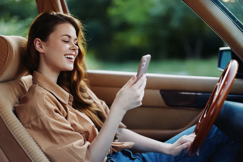 Young woman enjoying her smartphone inside a vintage car, smiling in a lush green background, wearing a casual brown shirt, showcasing a joyful mood.