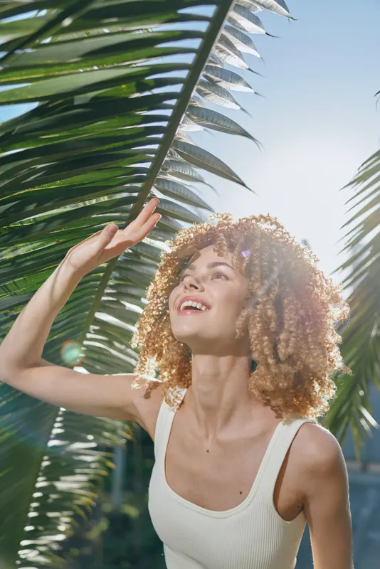 Smiling woman with curly hair enjoying bright sunlight under tropical palm leaves wearing white sleeveless top, radiating happiness and summer warmth outdoors.