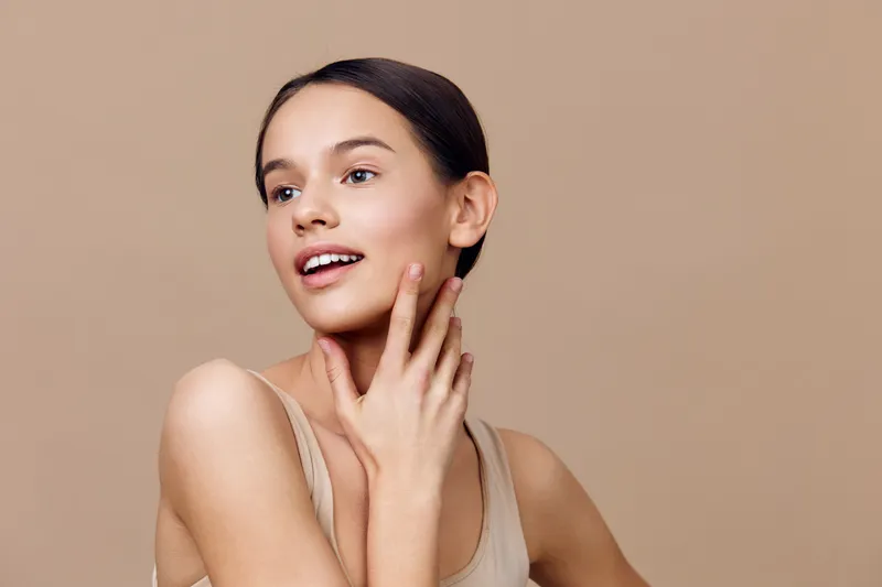 Young woman with a radiant smile showcases healthy skin in a minimalistic setting, wearing a neutral tank top, emphasizing natural beauty and confidence