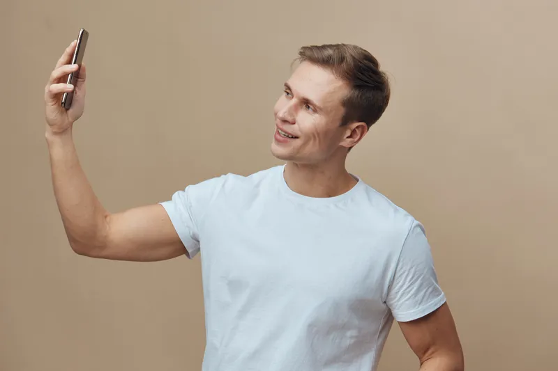 Smiling young man taking a selfie with smartphone on plain beige background. Casual white t-shirt, cheerful mood, modern technology, lifestyle concept, happiness, man in his 20s.
