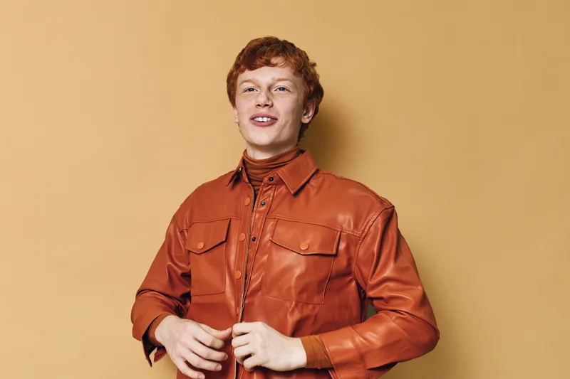 Young man with red hair wearing a brown jacket standing against beige background smiling confident caucasian male portrait casual fashion style