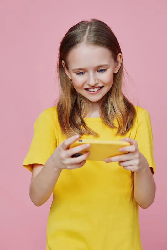 Happy girl with long hair in a yellow t shirt playing games on a smartphone against a pink background, expressing joy and excitement