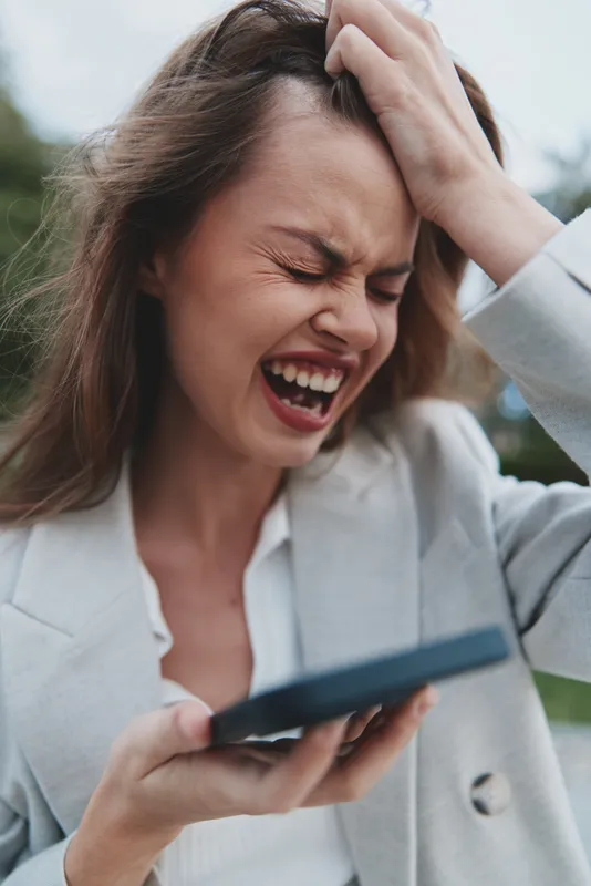 Frustrated woman in white blazer shouting on smartphone expressing stress and anxiety in outdoor natural setting with blurred green background