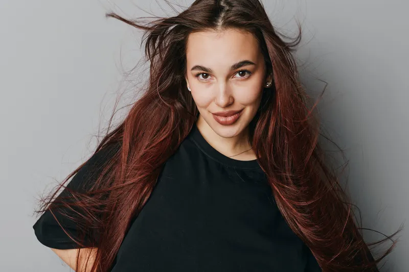 Young woman with long red hair wearing black shirt posing confidently against gray background with natural makeup and slight smile in studio portrait for fashion or beauty concept.