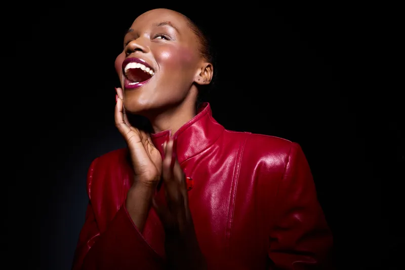 Smiling African woman with dark skin wearing a red leather jacket laughs joyfully against a black background. Concept of happiness, fashion, beauty, and confident emotions.
