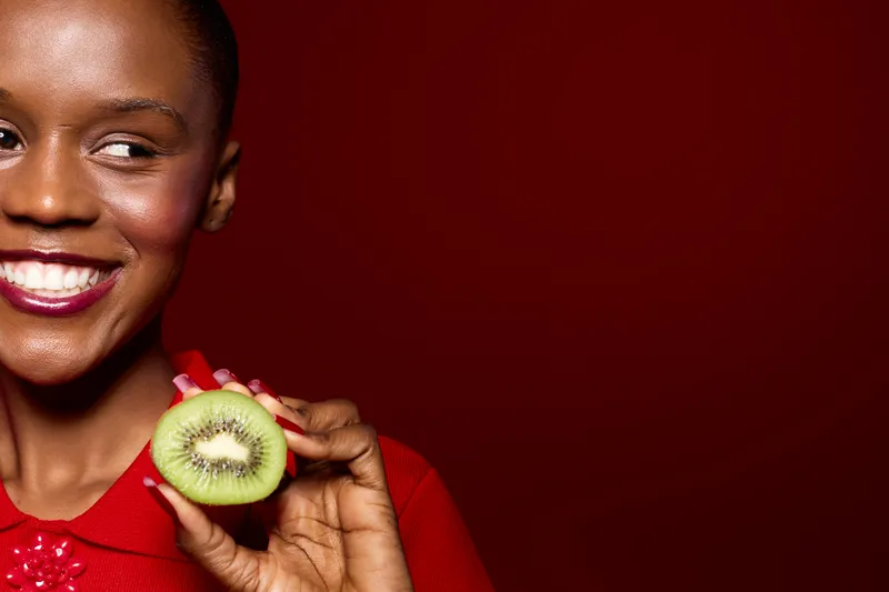 Happy African woman with dark skin smiles holding half of fresh kiwi fruit. Close-up on red background. Concept of healthy eating, natural food, wellness, and positive lifestyle.