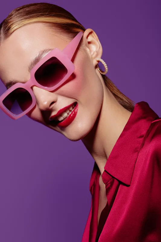 Stylish woman wearing oversized pink sunglasses and red lipstick smiles confidently against purple background. Fashion portrait with vibrant colors and modern accessories concept.